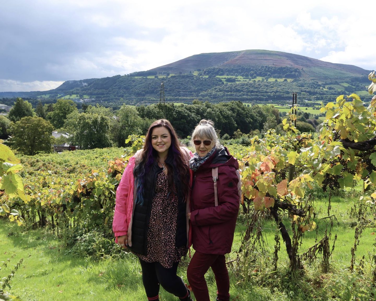 Luisa and her mum stand in the middle of Sugar Loaf Vineyard with the rolling Welsh hills behind them.