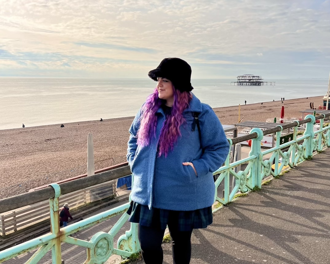 Luisa stands on the promenade on Brighton beach loooking out tot the sea with the old West Pier behind her. She wears a blue teddy bear jacket zipped up and a black furry hat
