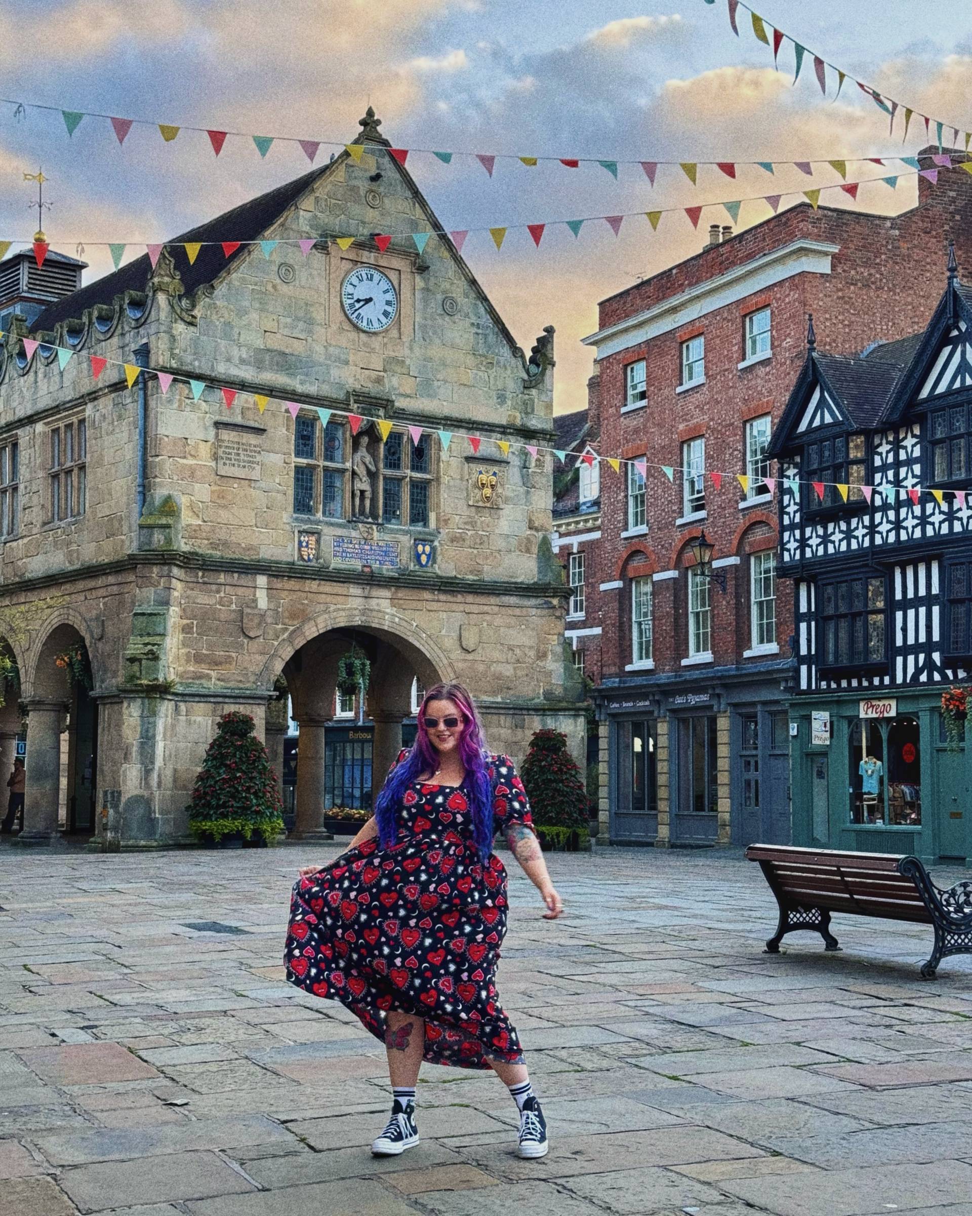 Luisa swooshes her black and red skirt in the Shrewsbury Town Square with a lovrly old building behind her, blue sky, and peach clouds and colourful bunting is in thr sky.